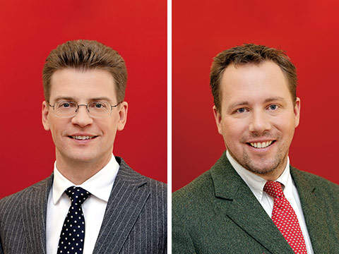 Zwei Männer in formeller Kleidung vor einem leuchtend roten Hintergrund. Der Mann links trägt eine Brille, einen grauen Nadelstreifenanzug und eine gepunktete Krawatte. Der Mann rechts hat einen Bart, trägt einen grünen Anzug und eine rote Krawatte. Beide lächeln.