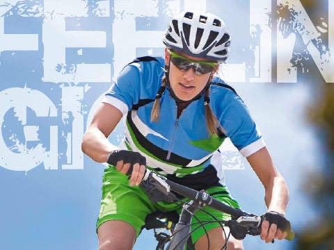 Ein Radfahrer in blau-grüner Kleidung mit Helm und Sonnenbrille fährt Fahrrad. Im Hintergrund ist ein großer, teilweise sichtbarer Text mit einem blauen Himmel dahinter zu sehen.