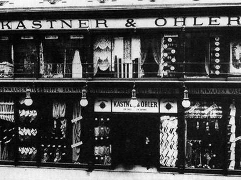 Schwarzweißfoto eines Gebäudes mit der Aufschrift „Kastner & Öhler“. Die Schaufensterfront zeigt verschiedene Stoffe und Textilien. Das Gebäude hat zwei Stockwerke und große Fenster präsentieren Produkte und dekorative Elemente.