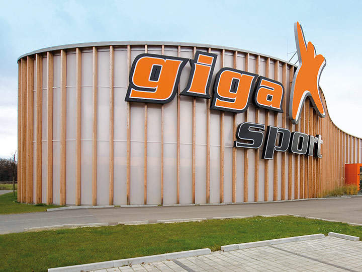Außenansicht eines großen, runden Gebäudes mit holzgetäfelter Fassade. Am Gebäude hängt ein großes orange-schwarzes Schild mit der Aufschrift „Gigasport“. Die Umgebung umfasst einen gepflasterten Weg und eine Rasenfläche.