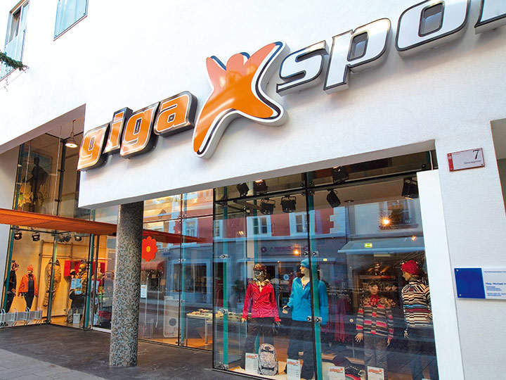 Straßenansicht eines Gigasport-Geschäfts mit moderner Fassade. Der Eingang ist von großen Glasfenstern flankiert, in denen Schaufensterpuppen in farbenfroher Sportbekleidung zu sehen sind. Über der Tür ist ein auffälliges „Gigasport“-Schild angebracht.