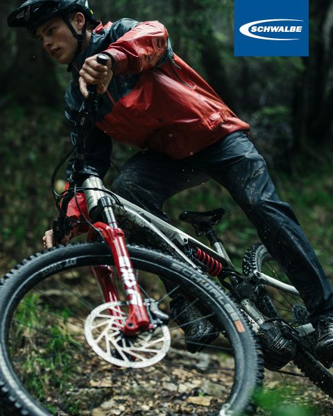 Ein Radfahrer in roter Jacke und Helm fährt mit seinem Mountainbike auf einem holprigen Trail. Das Rad hat eine rote Gabel und Scheibenbremsen. Der Hintergrund ist durch Grün verschwommen. Das Schwalbe-Logo ist in der oberen rechten Ecke sichtbar.