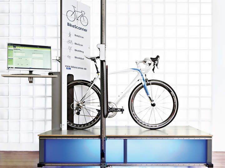 Ein weißes Rennrad mit blauen Akzenten auf einer blauen Plattform in einem Fahrradscanner-Setup. Links ist ein Computermonitor positioniert, der Daten anzeigt. Der Hintergrund zeigt ein Muster aus Milchglasblöcken.