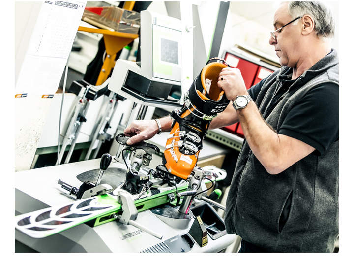 Ein Mann passt einen orangefarbenen Skischuh an einer Skibindungs-Einstellmaschine an. Er befindet sich in einer Werkstatt, im Hintergrund sind Werkzeuge und Geräte zu sehen. Er trägt eine Brille und eine schwarze Weste.