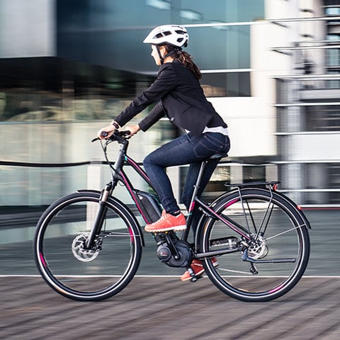 Eine Person mit Helm und Blazer fährt in einer städtischen Umgebung auf einem schwarz-rosa Fahrrad. Der Radfahrer ist in Bewegung und der Hintergrund ist verschwommen, was die Geschwindigkeit hervorhebt. Sie trägt Jeans und rote Turnschuhe.