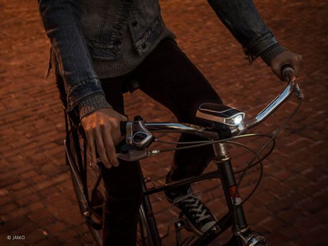 Eine Person in einer Jeansjacke fährt auf einem Vintage-Fahrrad mit schmalem Lenker über eine Kopfsteinpflasterstraße. Das Fahrrad hat ein leuchtendes Vorderlicht und ein schwarz-weißes Gerät, das am Lenker montiert ist.