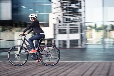 Eine Person mit Helm und Freizeitkleidung fährt mit einem Elektrofahrrad auf einem Holzweg. Im Hintergrund ist ein modernes Glasgebäude mit Spiegelungen und einer verschwommenen Stadtlandschaft zu sehen.