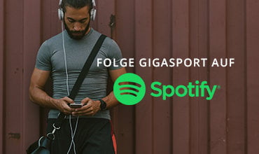 Ein Mann mit Kopfhörern und sportlichem Outfit schaut auf sein Handy. Der Text lautet „FOLGE GIGASPORT AUF Spotify“. Ein Spotify-Logo wird angezeigt. Der Hintergrund ist eine braune Wand.