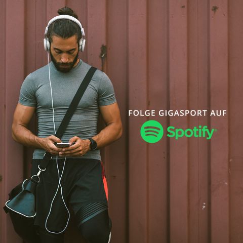 Ein Mann mit Kopfhörern lehnt an einer braunen Wand und schaut auf sein Handy. Er trägt ein graues Hemd, schwarze Shorts und hat eine Tasche über der Schulter. Darauf steht der Text „Folge Gigasport auf Spotify“ mit dem Spotify-Logo.