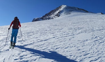Eine Person in roter Jacke und blauer Hose fährt unter einem klaren blauen Himmel einen schneebedeckten Hang hinauf auf einen Berggipfel. Auf dem Schnee sind Skispuren und lange Schatten zu sehen.