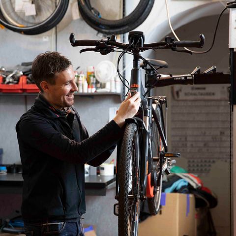 Eine Person lächelt, während sie in einer Werkstatt ein Fahrrad einstellt. Die Werkstatt ist voller Werkzeuge und Fahrradteile, und im Hintergrund hängen mehrere Räder an der Wand.