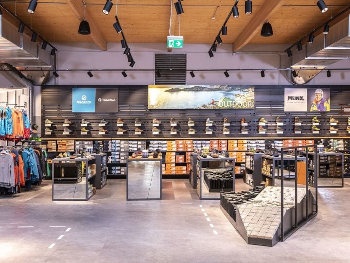 Ein modernes Schuhgeschäft mit einer Auswahl an Outdoor-Schuhen in Regalen. Marken sind auf einer Wandanzeige sichtbar. Das Geschäft verfügt über elegante Beleuchtung, verspiegelte Armaturen und einen Bereich zum Anprobieren von Schuhen auf einer strukturierten Rampe. Kleidung befindet sich auf der linken Seite.