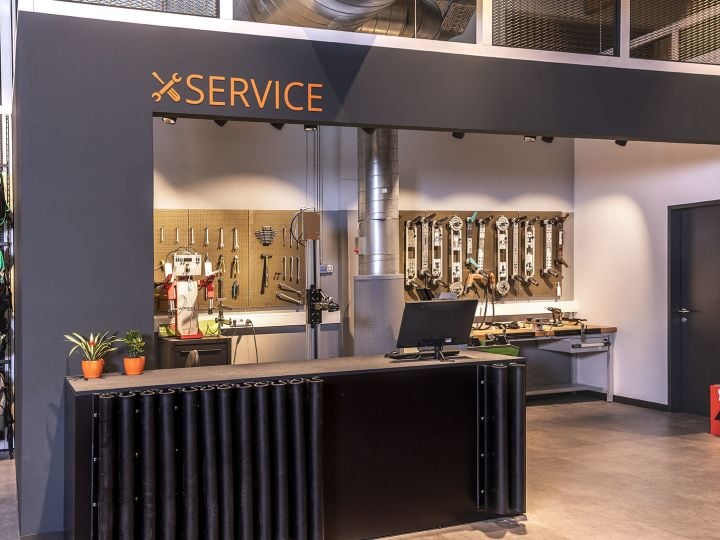 Skiservice-Shop mit Theke vorn, an der Wand montierten Werkzeugen und einer Arbeitsstation im Hintergrund. Links hängt die Skiausrüstung an Gestellen. Über der Theke prangt das Wort „SERVICE“.