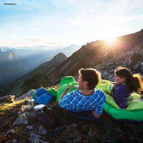 Ein Mann und eine Frau liegen in Schlafsäcken auf Isomatten und blicken auf die Berge bei Sonnenaufgang. Campingausrüstung, darunter Tassen und ein Rucksack, liegt in der Nähe. Der Himmel ist klar und wirft einen warmen Schein auf die Szene.