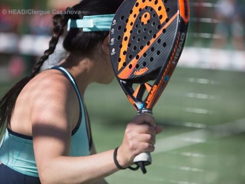 Eine Frau mit einem orange-schwarzen Schläger bereitet sich darauf vor, auf einem Padel-Platz einen Ball zu schlagen. Sie trägt ein blaues Tanktop und eine Mütze und steht mit dem Rücken zur Kamera. Im Hintergrund sind ein Netz und eine verschwommene Menschenmenge zu sehen.
