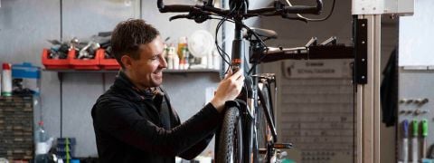 Ein Radfahrer mit Helm und Sonnenbrille fährt mit seinem Mountainbike auf einem Feldweg, der von leuchtenden Wildblumen und spärlichen Bäumen umgeben ist. Der Hintergrund zeigt eine raue, natürliche Landschaft unter einem klaren Himmel.
