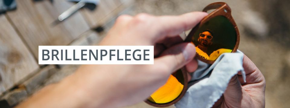 Eine Person putzt eine Sonnenbrille mit einem Tuch. In der Linse ist die Spiegelung einer Person zu sehen, die eine Sonnenbrille trägt. Das Wort „BRILLENPFLEGE“ ist über das Bild gelegt. Der Hintergrund zeigt eine Holzoberfläche.