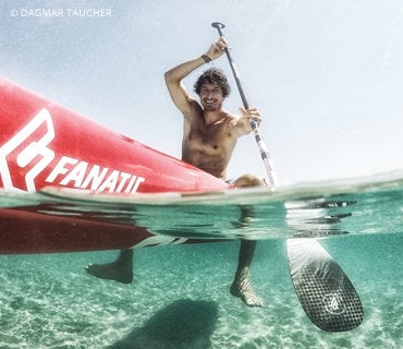 Ein Mann ohne Hemd sitzt auf einem roten Paddleboard in klarem, seichtem Wasser und hält in einer Hand ein Paddel. Das Foto ist teilweise unter Wasser und zeigt das Board und den Sandboden. Der Text auf dem Board lautet „Fanatic“.