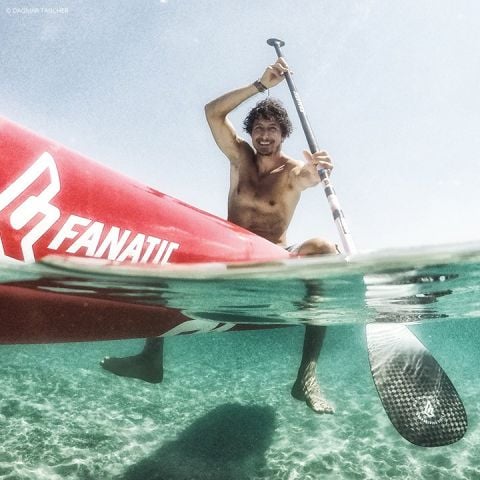 Eine Person paddelt auf einem roten Stand-Up-Paddleboard über klares blaues Wasser. Das Bild wurde halb unter Wasser aufgenommen und zeigt sowohl den Paddler als auch den unter Wasser liegenden Teil des Boards und des Paddels. Die Person scheint zu lächeln und die Aktivität zu genießen.