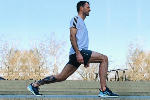 Ein Mann macht im Freien auf einem Steinboden eine Ausfallschrittübung. Er trägt ein weißes T-Shirt, schwarze Shorts und Turnschuhe. Im Hintergrund sind blattlose Bäume und ein klarer Himmel zu sehen. Auf seinem rechten Bein ist eine Tätowierung zu sehen.