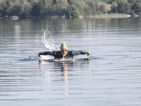 Eine Person in schwarzem Neoprenanzug und Schwimmbrille schwimmt im offenen Wasser und führt den Schmetterlingsstil aus. Um sie herum sind Spritzer zu sehen. Bäume säumen das entfernte Ufer unter einem klaren Himmel.