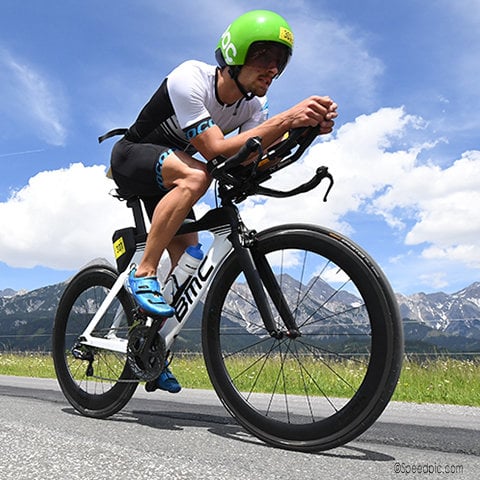 Ein Radfahrer mit grünem Helm und schwarz-weißer Kleidung fährt auf einem BMC-Fahrrad auf einer asphaltierten Straße. Im Hintergrund ist eine malerische Aussicht auf Berge unter einem blauen Himmel mit vereinzelten Wolken zu sehen.