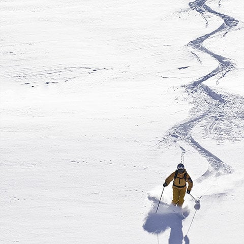 Ein Skifahrer in gelbem Outfit gleitet einen schneebedeckten Hang hinunter und hinterlässt eine gewundene Spur. Die Szene ist hell von unberührtem Schnee und der Skifahrer wirft einen Schatten auf die weiße Oberfläche.