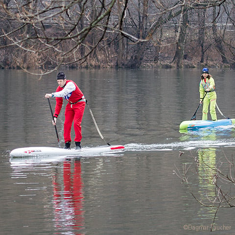 Zwei Personen fahren Stand-Up-Paddleboard auf einem ruhigen Fluss. Die Person vorne trägt ein rotes Outfit und einen roten Hut, während die Person dahinter ein hellgrünes Outfit und einen Helm trägt. Im Hintergrund säumen kahle Bäume das Flussufer.