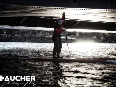 Eine Person, die auf einem Fluss unter einer Brücke paddelt, trägt Outdoor-Ausrüstung und ein Stirnband. Die Beleuchtung erzeugt einen dramatischen Kontrast zum Wasser und unter der Brücke. Das Logo „AUCHER Photography“ ist in der Ecke sichtbar.