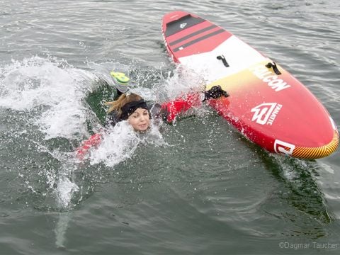 Person in roter Jacke fällt neben einem umgekippten roten Paddleboard ins Wasser. Sie hat einen Fuß in der Luft und das Wasser spritzt herum.