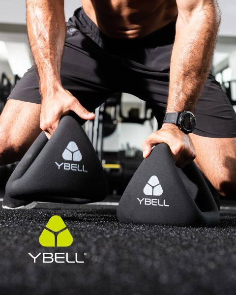 Eine Person in Trainingskleidung hält zwei schwarze YBELL-Gewichte auf dem Boden des Fitnessstudios. Sie trägt eine Uhr und Shorts. Das YBELL-Logo ist auf den Gewichten und in der unteren linken Ecke abgebildet.