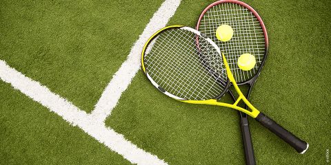 Zwei Tennisschläger und Bälle auf einem grünen Rasenplatz, in der Nähe weißer Begrenzungslinien positioniert. Die Schläger sind gekreuzt, wobei einer einen gelben Rahmen und der andere einen schwarzen Rahmen hat.