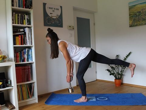 Eine Person balanciert auf einem Bein, hält in der rechten Hand eine Hantel und führt auf einer blauen Yogamatte ein einbeiniges Kreuzheben aus. Ein Bücherregal, eine Topfpflanze und Wandkunst schmücken den Raum.