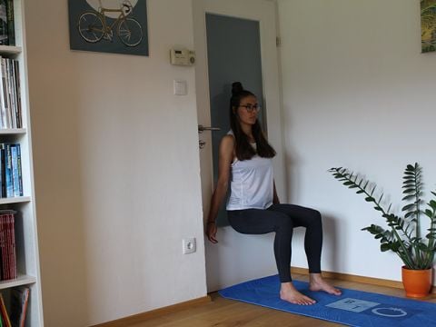 Eine Person führt in einem Raum eine Wandsitzübung durch. Sie steht mit dem Rücken an einer Tür, die Knie sind im 90-Grad-Winkel gebeugt. Unter ihr liegt eine blaue Yogamatte, daneben steht eine Topfpflanze. Bücherregale und Wandkunst sind sichtbar.