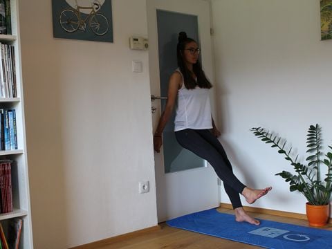 Eine Person in einem weißen Tanktop und schwarzen Leggings lehnt an einer Tür und macht eine Einbeinübung. Sie steht auf einer blauen Trainingsmatte, daneben steht eine Pflanze. Im Hintergrund sind ein Bücherregal und ein Fahrradbild zu sehen.