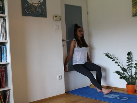 Person, die in einem Raum eine Wandsitzübung macht. Sie trägt ein weißes Tanktop und schwarze Leggings. Sie lehnt an einer weißen Tür, unter der sich eine blaue Trainingsmatte befindet. Im Hintergrund sind ein Bücherregal, eine Pflanze und Wandkunst zu sehen.