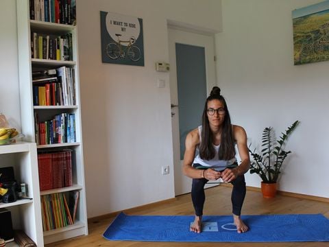 Eine Person mit langen Haaren und Brille hockt auf einer blauen Gymnastikmatte in einem Raum mit Holzboden. Im Hintergrund sind Bücherregale, eine Topfpflanze und ein Wandbild mit einem Fahrrad zu sehen.
