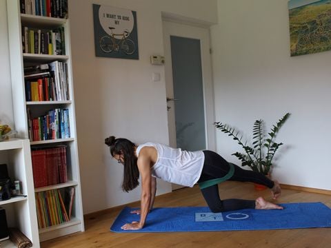 Eine Person trainiert drinnen auf einer blauen Yogamatte. Sie trägt ein weißes Tanktop und schwarze Leggings. Sie befindet sich in einer knienden Plank-Position mit einem ausgestreckten Bein. Im Raum stehen ein Bücherregal, eine Tür und im Hintergrund eine Pflanze.