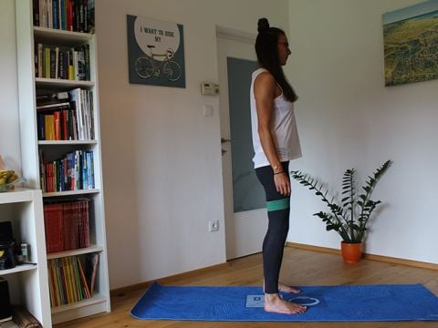 Eine Person steht in einem Raum auf einer blauen Yogamatte. Sie trägt ein weißes Tanktop und schwarze Leggings mit einem grünen Widerstandsband um die Oberschenkel. Im Hintergrund sind ein Bücherregal, ein Poster mit Fahrradmotiv und eine Pflanze zu sehen.