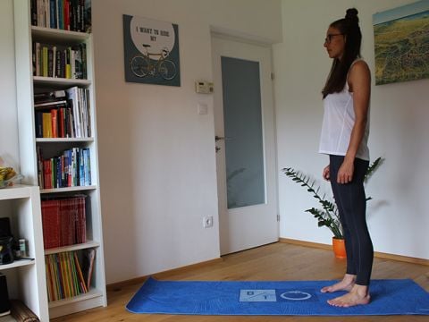Eine Frau steht auf einer blauen Yogamatte in einem Raum. Sie trägt ein weißes ärmelloses Oberteil und schwarze Leggings. Hinter ihr steht ein Bücherregal mit Büchern, einem Wandposter mit einem Fahrrad, einer Tür und einer Topfpflanze.