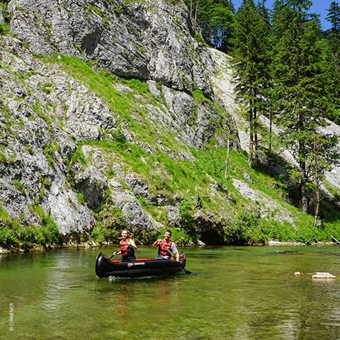 Zwei Menschen mit Schwimmwesten paddeln in einem Kanu auf einem klaren, seichten Fluss. Sie sind umgeben von üppigem Grün, felsigen Klippen und hohen Bäumen unter einem strahlend blauen Himmel.