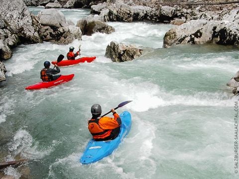 Drei Kajakfahrer befahren einen felsigen Wildwasserfluss. Sie tragen Helme und Schwimmwesten. Die Kajaks sind rot, blau und orange. Das Wasser ist schnell und die umliegenden Felsen sind schroff. Die Szene suggeriert Abenteuer und Teamwork.