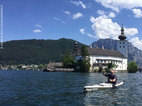Eine Person fährt Kajak auf einem ruhigen See vor der Kulisse eines malerischen Gebäudes mit Kuppel und Türmen. Ein üppig grüner Berg steht unter einem klaren blauen Himmel und das Wasser spiegelt die lebendige Szene wider.