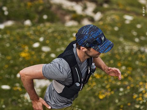Eine Person läuft im Freien auf einer Wiese. Sie trägt eine blau gemusterte Mütze, ein graues T-Shirt und eine schwarze Trinkweste. Das Gelände ist mit gelben Blumen und Steinen übersät. Der Läufer ist konzentriert und bewegt sich zügig.