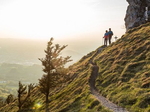 Ein Paar steht bei Sonnenaufgang oder Sonnenuntergang auf einem schmalen Pfad entlang eines grasbewachsenen Hügels und blickt auf ein Tal. In der Nähe stehen ein einsamer Baum und Felsformationen, und warmes Sonnenlicht erhellt die Landschaft.