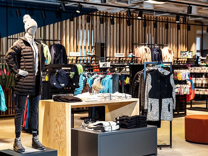Innenansicht eines Bekleidungsgeschäfts mit einer Schaufensterpuppe in schwarzer Jacke, weißem Hut und Stiefeln. Verschiedene bunte Jacken und Sportbekleidung sind auf Ständern und Regalen ausgestellt, mit Holzakzenten und moderner Beleuchtung im Hintergrund.