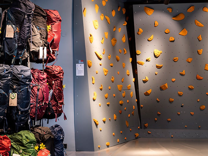 Indoor-Kletterwand mit verschiedenen orangefarbenen Griffen auf grauem Untergrund. Links daneben sind auf Gestellen mehrere Rucksäcke in unterschiedlichen Farben ausgestellt. Die Beleuchtung ist hell und der Boden poliert.