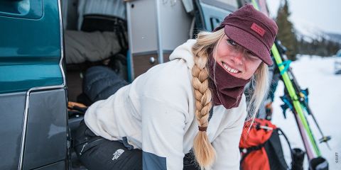Eine Person mit langem Zopf und kastanienbrauner Mütze lächelt, während sie aus einem Lieferwagen steigt. Sie ist warm angezogen, von Schnee umgeben, und draußen sind Skier und Schneeausrüstung zu sehen.