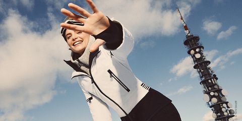 Eine Person in Winterkleidung streckt ihre Hand spielerisch in Richtung Kamera, während sie vor einem blauen Himmel mit Wolken steht. Auf der rechten Seite ist ein hoher Kommunikationsturm zu sehen.