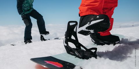 Nahaufnahme von zwei Personen, die auf einem schneebedeckten Berghang Snowboard fahren. Einer trägt leuchtend orangefarbene Hosen und schwarze Stiefel und steht neben einem Snowboard. Der andere in grün-schwarzer Kleidung ist im Hintergrund. Der Himmel ist klar und blau.
